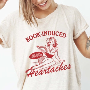 May include: A white t-shirt with red text that reads "BOOK-INDUCED HEARTACHES" and a cartoon image of a woman crying while reading a book. The text in a speech bubble above the woman reads "THE PLOT TWIST IS PAIN."