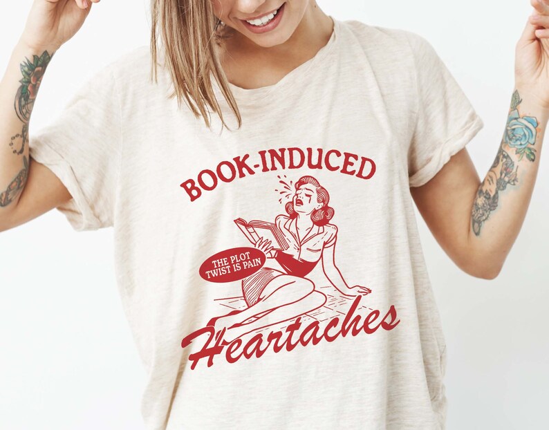 May include: A white t-shirt with red text that reads "BOOK-INDUCED HEARTACHES" and a cartoon image of a woman crying while reading a book. The text in a speech bubble above the woman reads "THE PLOT TWIST IS PAIN."