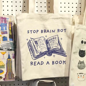 May include: A white tote bag with a blue and white illustration of an open book and the text "STOP BRAIN ROT READ A BOOK".