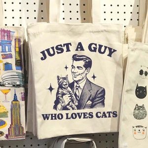 May include: A white canvas tote bag with a blue graphic of a man holding a cat. The graphic says "Just a guy who loves cats".