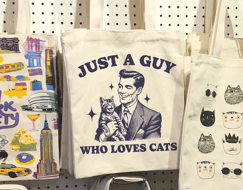 May include: A white canvas tote bag with a blue graphic of a man holding a cat. The graphic says "Just a guy who loves cats".