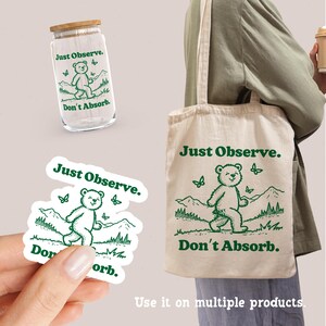 May include: A white tote bag with green text that reads "Just Observe. Don't Absorb." and a green illustration of a bear walking. The bag has green butterfly illustrations and green mountain illustrations.