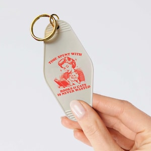 May include: White motel key chain with a red illustration of a woman reading a book with a cat in her lap. The text reads "Time spent with books & cats is never wasted."