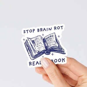 May include: White sticker with blue text and illustration. The text reads "Stop Brain Rot Read a Book". The illustration is of an open book with a blue outline.