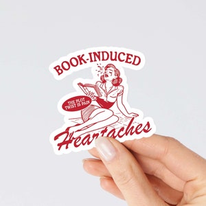 May include: Red and white sticker with a cartoon woman crying and reading a book. The sticker says "Book-Induced Heartaches" and "The plot twist is pain".