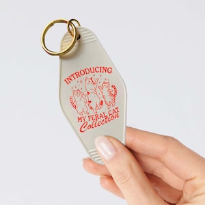 Puede incluir: Llavero de motel blanco con un dise&ntilde;o rojo que presenta tres gatos bailando y el texto "Introducing My Feral Cat Collection".