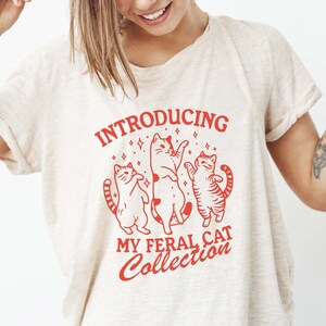 Puede incluir: Una camiseta blanca con un dise&ntilde;o gr&aacute;fico rojo que dice "Introducing My Feral Cat Collection". El gr&aacute;fico presenta tres gatos estilizados bailando con estrellas a su alrededor.