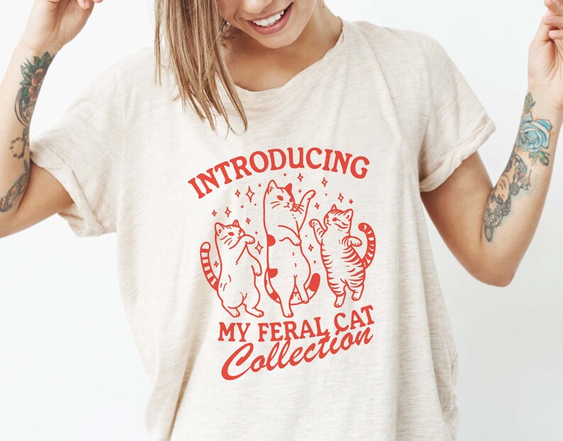 Puede incluir: Una camiseta blanca con un dise&ntilde;o gr&aacute;fico rojo que dice "Introducing My Feral Cat Collection". El gr&aacute;fico presenta tres gatos estilizados bailando con estrellas a su alrededor.