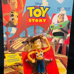 K&ouml;nnte beinhalten: Sammelfigur von Woody aus Toy Story, eine Cowboy-Puppe mit braunem Hut, gelbem Hemd, blauer Jeans und rotem Halstuch. Die Figur steht auf einem sternf&ouml;rmigen Sockel. Die Verpackung ist rot und blau mit dem Toy Story-Logo und dem Text "Ages 3 and Up".