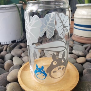 May include: A clear glass cup with a white and grey leaf design and blue and grey cartoon characters. The cup is sitting on a wooden coaster.