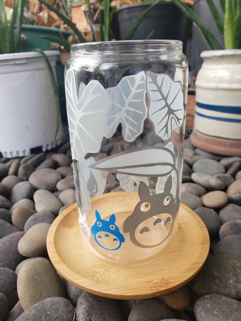 May include: A clear glass cup with a white and grey leaf design and blue and grey cartoon characters. The cup is sitting on a wooden coaster.