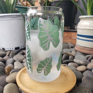 May include: A clear glass can with a green leaf design. The can is filled with a white liquid and is sitting on a wooden coaster. The coaster is on a bed of grey stones.