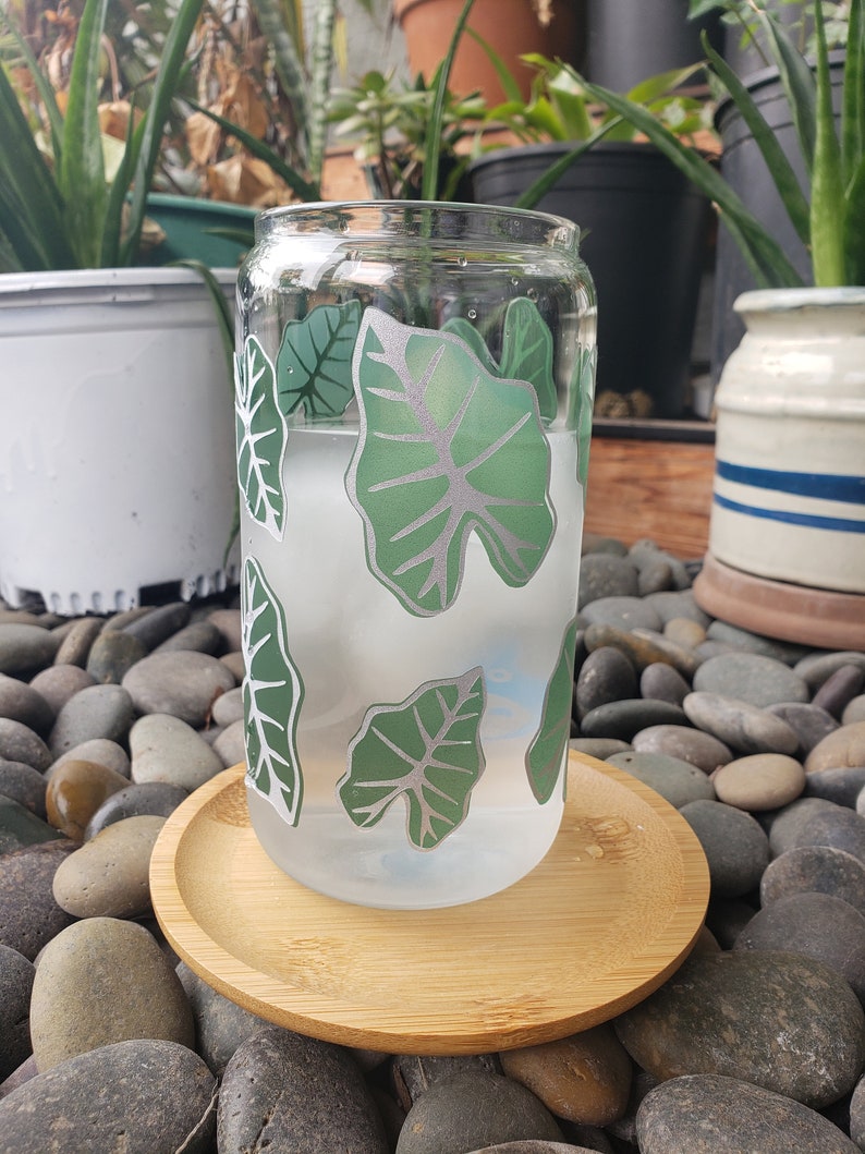 May include: A clear glass can with a green leaf design. The can is filled with a white liquid and is sitting on a wooden coaster. The coaster is on a bed of grey stones.
