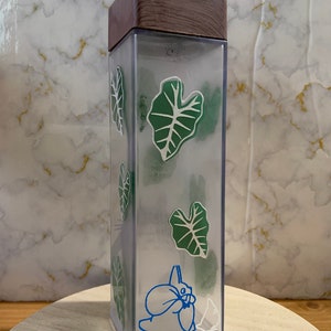 May include: A clear plastic water bottle with a brown wooden lid. The bottle features a green leaf design and a blue cartoon character on the side.