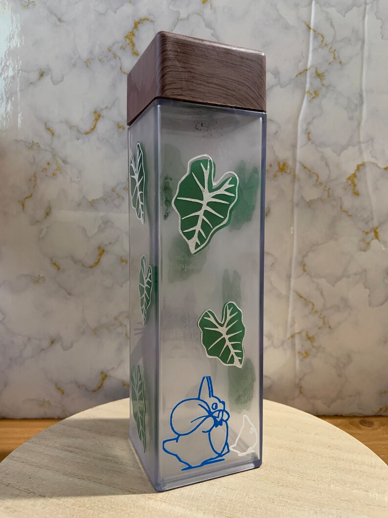 May include: A clear plastic water bottle with a brown wooden lid. The bottle features a green leaf design and a blue cartoon character on the side.