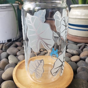 May include: A clear glass drinking glass with a white leafy design and a blue cartoon character. The glass is sitting on a wooden coaster.