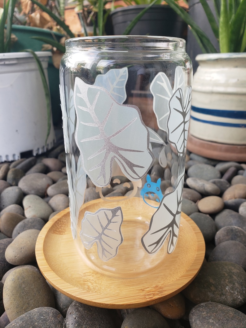 May include: A clear glass drinking glass with a white leafy design and a blue cartoon character. The glass is sitting on a wooden coaster.