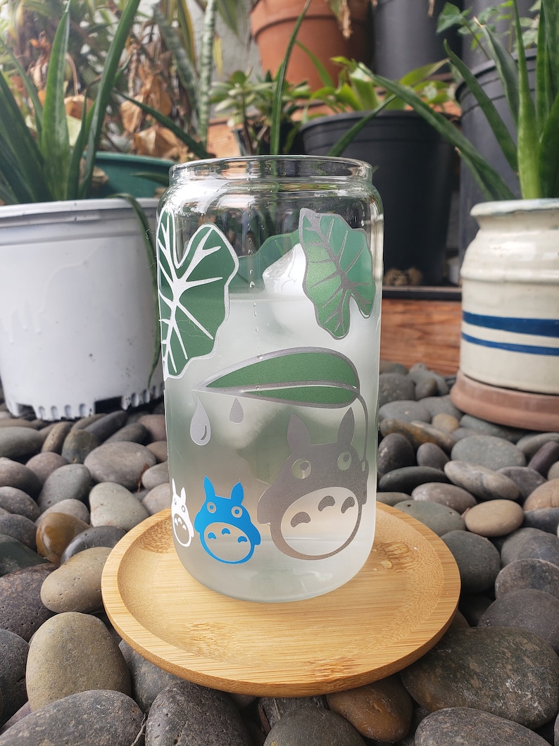 May include: A clear glass drinking glass with a green and white leafy design and blue cartoon characters. The glass is sitting on a wooden coaster.