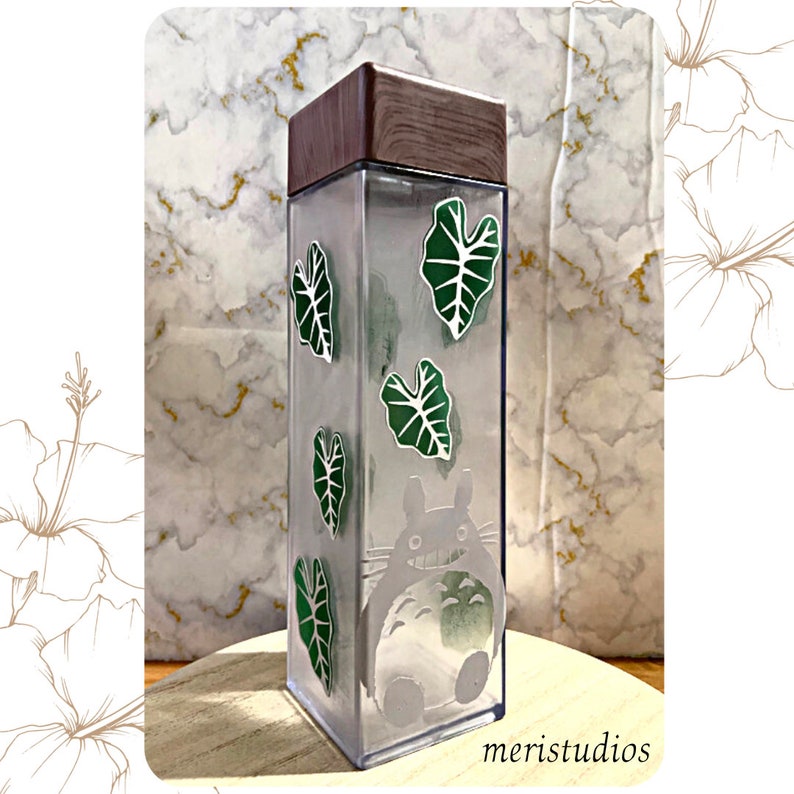 May include: A clear plastic water bottle with a brown wooden lid. The bottle features a graphic of a cartoon creature and green leaves.