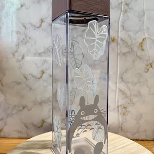 May include: A clear plastic water bottle with a brown wooden lid. The bottle features a white graphic of a cartoon character and leaves.