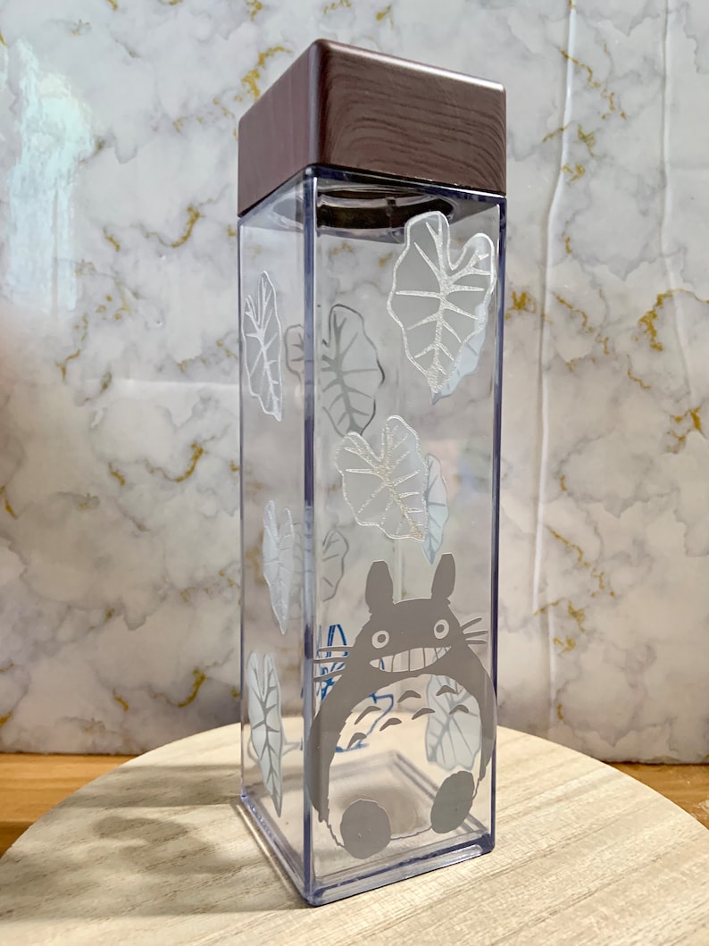 May include: A clear plastic water bottle with a brown wooden lid. The bottle features a white graphic of a cartoon character and leaves.