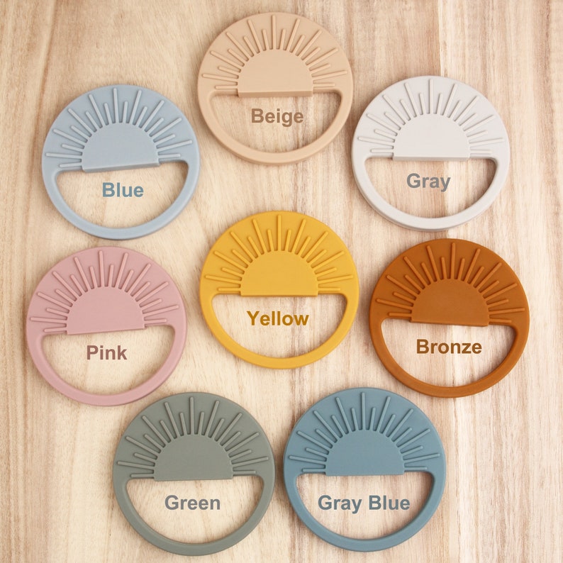 On the wooden table, there are eight personalized silicone baby soothing teether cups with different colors. The colors are as follows: gray; blue; yellow; pink; beige; bronze; green; gray-blue.