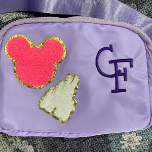 Op de afbeelding: Een paarse stoffen tas met een roze Mickey Mouse-hoofd en een witte hand met gouden glitter. De tas heeft een paarse band en de letters "GF" zijn geborduurd op de voorkant.