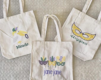 May include: Three white canvas tote bags with embroidered designs. The first bag has a pelican with a jester's hat and the name "Meade" embroidered on it. The second bag has three Mardi Gras masks and the name "jane jane" embroidered on it. The third bag has a Mardi Gras mask and the name "Higgins" embroidered on it.