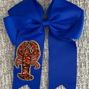 May include: A blue hair bow with a red and gold lobster design. The lobster is made of sequins and has a gold outline.