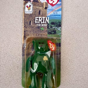 Può includere: Un orsacchiotto di peluche verde di nome "Erin" con un trifoglio sul petto, confezionato in una custodia di plastica trasparente. La confezione presenta un'illustrazione di un castello e il testo "Collect All 4!" e "Erin The Bear". Un'etichetta rossa a forma di cuore è attaccata.