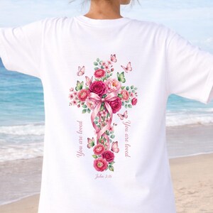 Puede incluir: Camiseta blanca con un dise&ntilde;o de cruz floral. La cruz est&aacute; adornada con rosas rosas, un lazo rosa y mariposas. Las palabras "You are loved" y "John 3:16" est&aacute;n impresas en la camiseta. El dise&ntilde;o est&aacute; en la espalda.