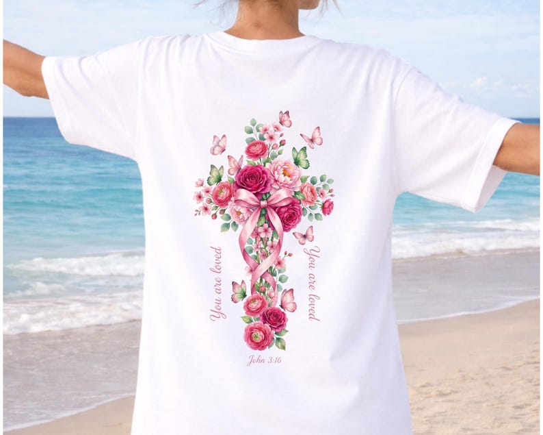 Puede incluir: Camiseta blanca con un dise&ntilde;o de cruz floral. La cruz est&aacute; adornada con rosas rosas, un lazo rosa y mariposas. Las palabras "You are loved" y "John 3:16" est&aacute;n impresas en la camiseta. El dise&ntilde;o est&aacute; en la espalda.