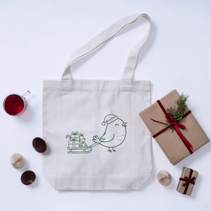 May include: A white canvas tote bag with a green outline illustration of a bird wearing a Santa hat pulling a sled of gifts. A wrapped gift with a red ribbon and greenery, a glass of red liquid, and paper decorations are also present.