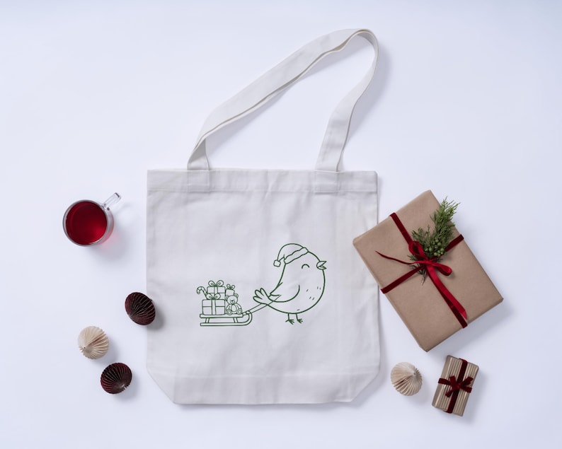 May include: A white canvas tote bag with a green outline illustration of a bird wearing a Santa hat pulling a sled of gifts. A wrapped gift with a red ribbon and greenery, a glass of red liquid, and paper decorations are also present.