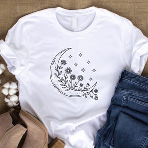 May include: White t-shirt featuring a black line drawing of a crescent moon with floral and star details. The shirt is paired with blue denim jeans and tan ankle boots, creating a casual, everyday aesthetic.