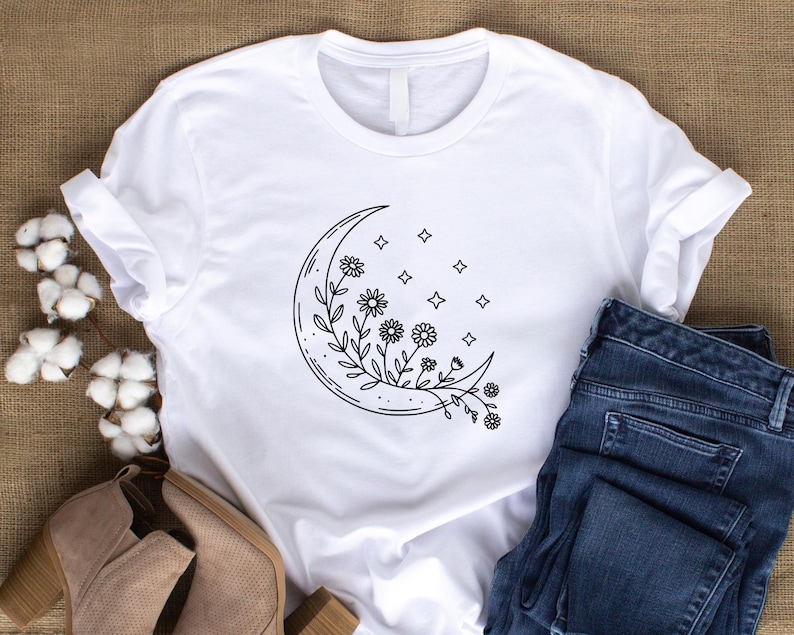 May include: White t-shirt featuring a black line drawing of a crescent moon with floral and star details. The shirt is paired with blue denim jeans and tan ankle boots, creating a casual, everyday aesthetic.