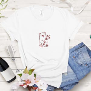 May include: White t-shirt featuring a cartoon bear holding a candy cane. The bear and candy cane are in a reddish-brown color. The shirt is paired with blue denim shorts. A bottle of champagne, a champagne flute, and sunglasses are also in the image.