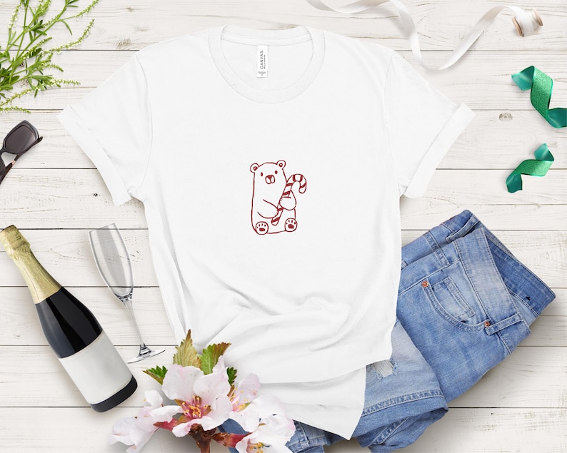 May include: White t-shirt featuring a cartoon bear holding a candy cane. The bear and candy cane are in a reddish-brown color. The shirt is paired with blue denim shorts. A bottle of champagne, a champagne flute, and sunglasses are also in the image.