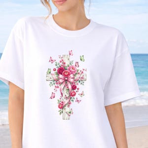 Puede incluir: Camiseta blanca con un dise&ntilde;o de acuarela de una cruz de madera adornada con rosas rosas, un lazo rosa y mariposas. La cruz es el punto central, con el arreglo floral y las mariposas que a&ntilde;aden un toque de elegancia. El dise&ntilde;o est&aacute; sobre un fondo blanco.