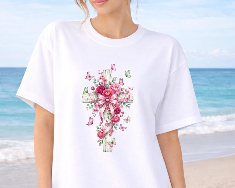 Puede incluir: Camiseta blanca con un dise&ntilde;o de acuarela de una cruz de madera adornada con rosas rosas, un lazo rosa y mariposas. La cruz es el punto central, con el arreglo floral y las mariposas que a&ntilde;aden un toque de elegancia. El dise&ntilde;o est&aacute; sobre un fondo blanco.
