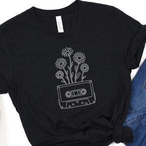 May include: Black t-shirt featuring a white line drawing of daisies growing out of a cassette tape. The shirt has a crew neck and short sleeves. The tag on the shirt reads "Canvas". The shirt is paired with blue jeans and a leopard print shoe.