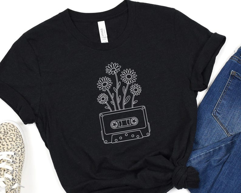 May include: Black t-shirt featuring a white line drawing of daisies growing out of a cassette tape. The shirt has a crew neck and short sleeves. The tag on the shirt reads "Canvas". The shirt is paired with blue jeans and a leopard print shoe.