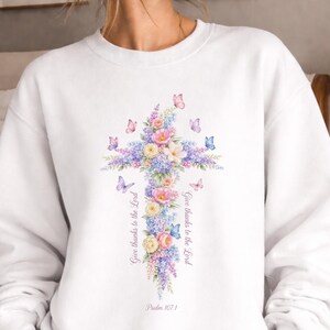 Puede incluir: Sudadera blanca con un dise&ntilde;o de cruz floral con flores y mariposas en colores pastel. La cruz est&aacute; adornada con flores rosas, moradas y azules. El texto "Give thanks to the Lord" y "Psalm 107:1" est&aacute; impreso en la sudadera.