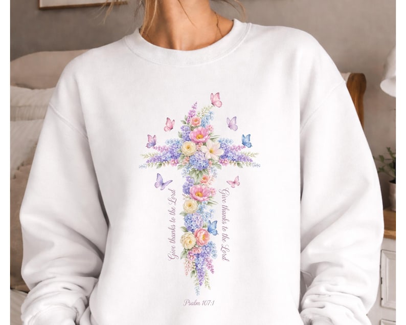 Puede incluir: Sudadera blanca con un dise&ntilde;o de cruz floral con flores y mariposas en colores pastel. La cruz est&aacute; adornada con flores rosas, moradas y azules. El texto "Give thanks to the Lord" y "Psalm 107:1" est&aacute; impreso en la sudadera.