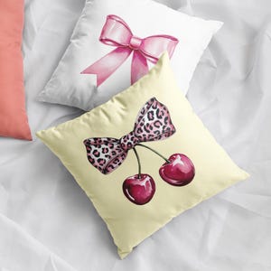 May include: A yellow pillow with a pink and leopard print bow and two red cherries. The pillow is on a white surface.