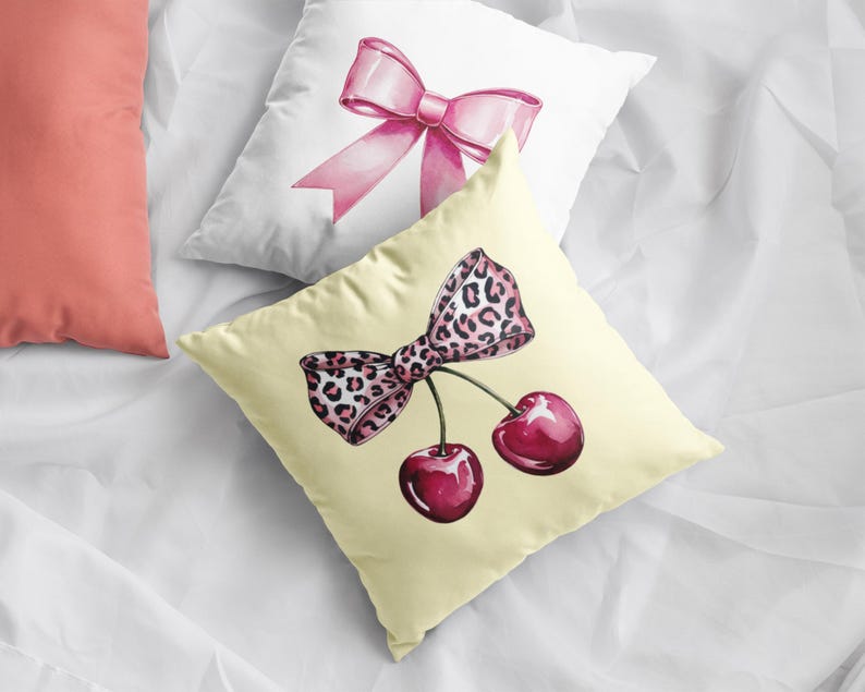 May include: A yellow pillow with a pink and leopard print bow and two red cherries. The pillow is on a white surface.