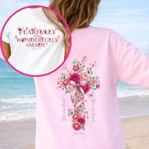 Puede incluir: Una sudadera rosa claro con un diseño de cruz floral en la espalda. La cruz está adornada con rosas rosas, vegetación y mariposas. El texto "You are loved" y "John 3:16" están impresos en la sudadera. Una camiseta blanca con el texto "FEARFULLY AND WONDERFULLY MADE" es visible.