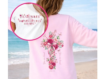 Puede incluir: Una sudadera rosa claro con un diseño de cruz floral en la espalda. La cruz está adornada con rosas rosas, vegetación y mariposas. El texto "You are loved" y "John 3:16" están impresos en la sudadera. Una camiseta blanca con el texto "FEARFULLY AND WONDERFULLY MADE" es visible.