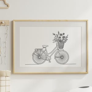 May include: A framed black and white line drawing of a bicycle with a basket of flowers. The bicycle has a classic design with a rear rack and is set against a white background. The frame is a light wood color.