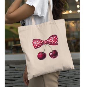 May include: A white canvas tote bag with a red and white polka dot bow and two red cherries. The cherries have stems and leaves.